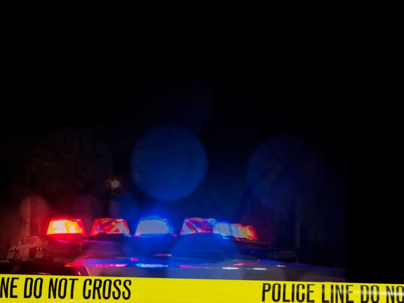 A police car with flashing lights behind yellow tape that reads: "Police line, do not cross.”