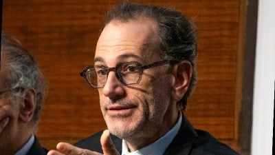 A middle-aged man in glasses gestures while speaking, wearing a suit and tie, with a wooden background visible.