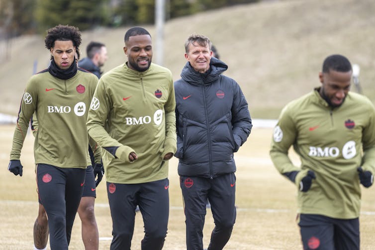 A middle-aged white man in a parka walks along a field, flanked by three Black soccer players in olive green long-sleeves