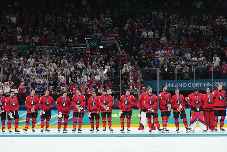 A row of men in red hockey uniforms stand in line on the ice wearing silver medals