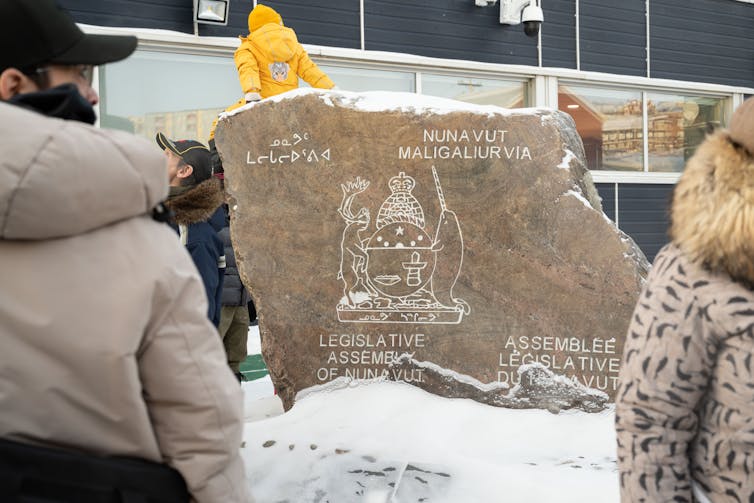 People stand facing a rock carved with the words Legislative Assembly of Nunavut