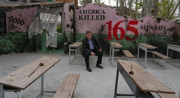A man sits on a bench. Behind him a mural reads 'America killed 165.'