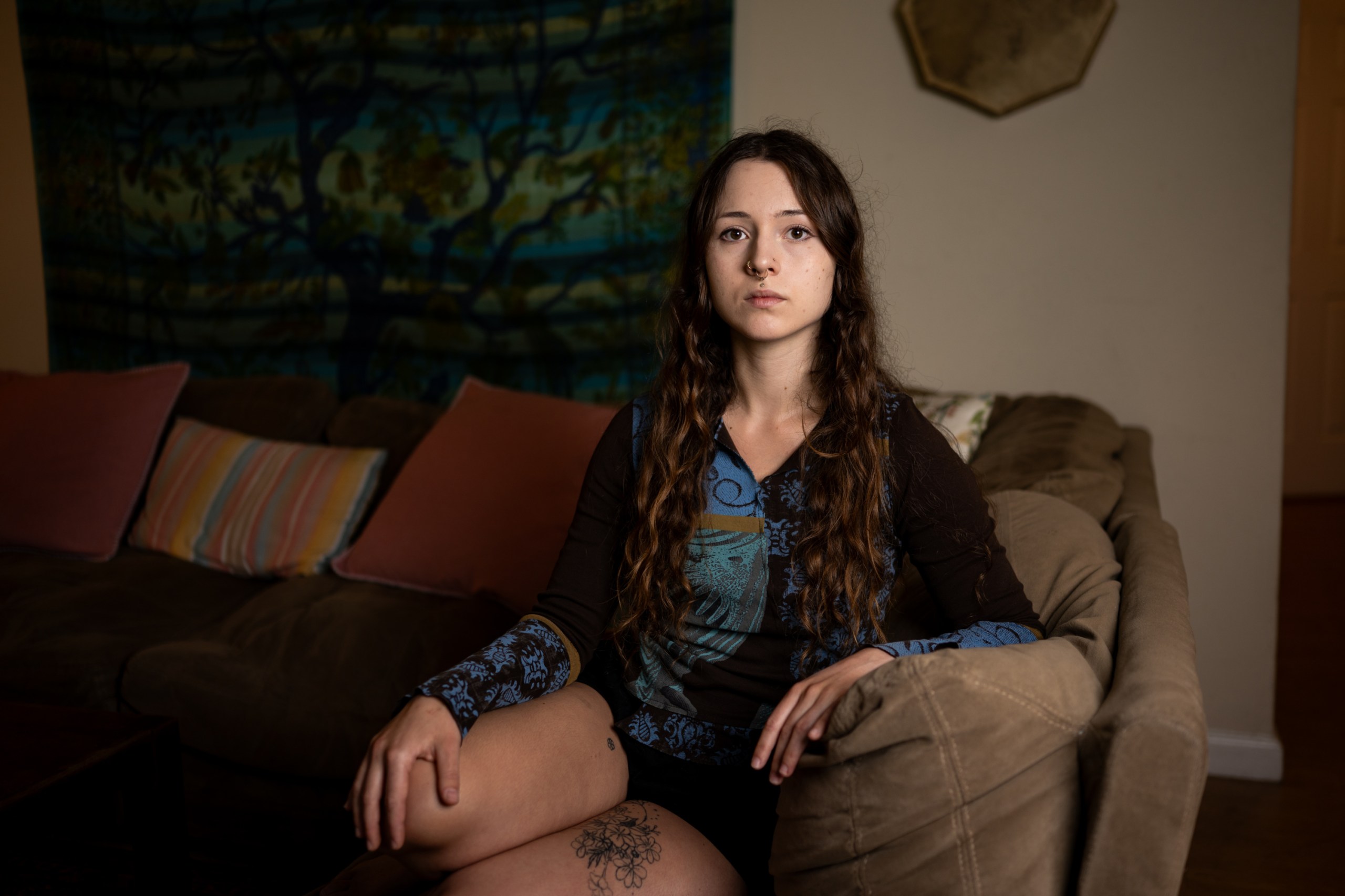A woman in her 20s sits on a sofa and looks at the camera with a serious expression.