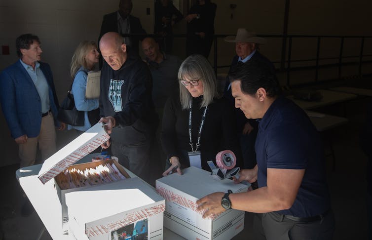 A man in a hoodie with carboard boxes on a table next to two people and a crowd behind.