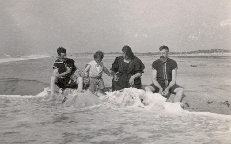 A family in old fashioned long bathing suits sit in the sea