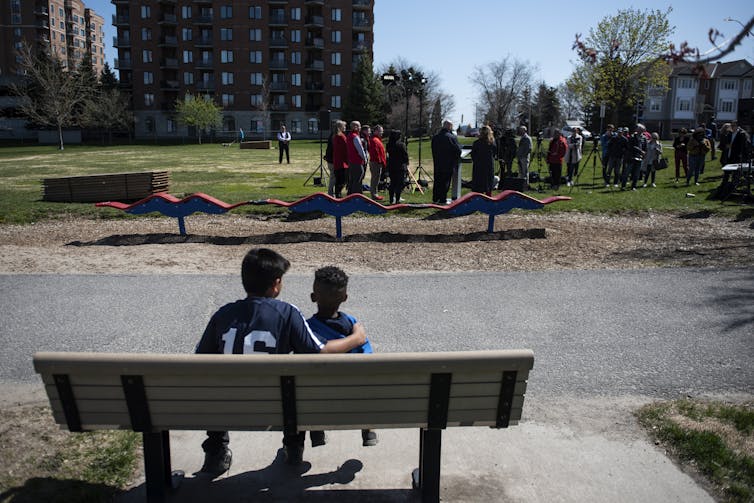 An older child seen with their arm draped around a younger child, sitting on a park bench and gazing at adults in a park and a play structure.