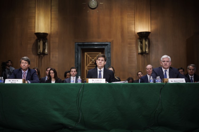 Three men wearing suits sit at a green table with people sitting behind them in a wood paneled room.