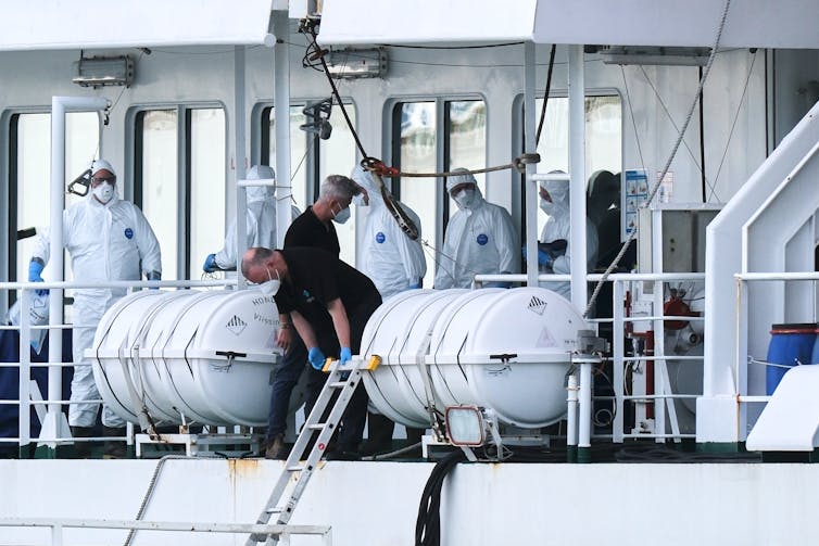 People in white protective suits on the deck of a boat with two people wearing dark shirts and face masks