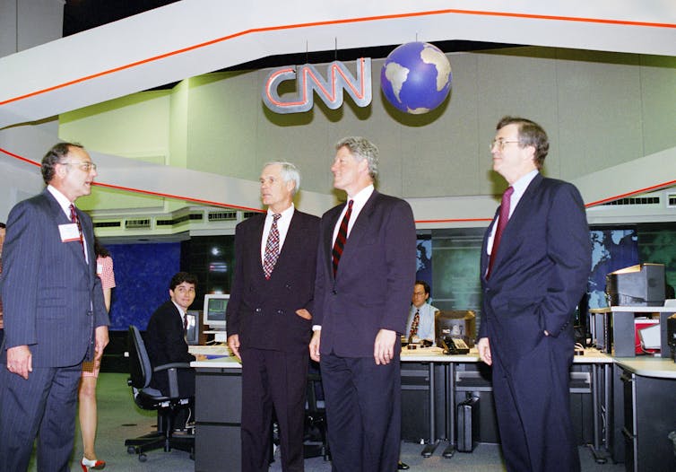 Four men dressed in suits stand in a newsroom.