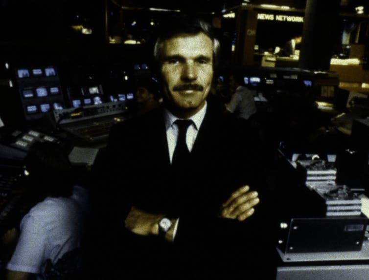 A man stands in a newsroom, arms folded.