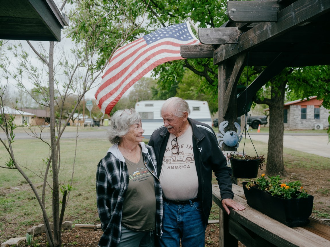 An older woman and man, wearing jeans and jackets and smiling at each other, stand in a grassy area near some houses. An American flag blows in the wind behind them.