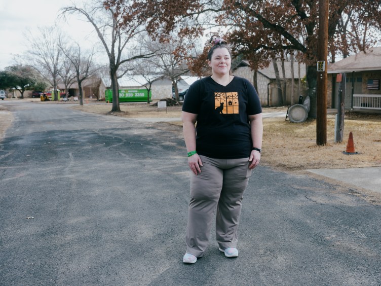 A woman wearing a black T-shirt with a yellow graphic and gray pants stands in a road through a residential neighborhood.