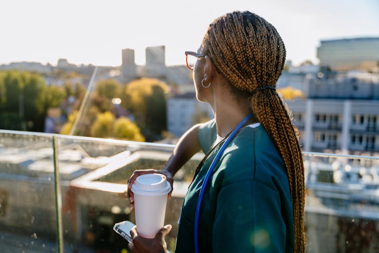 female healthcare worker on a break outside on a hospital balcony with a coffee and her phone in her hand.