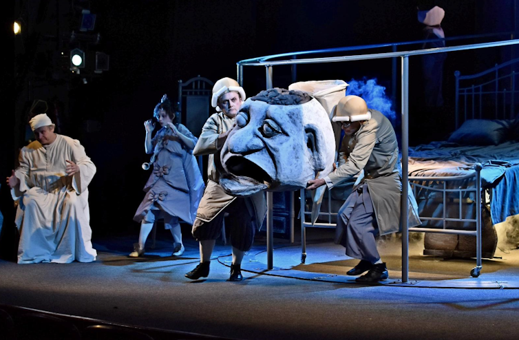 A theatre troupe on stage with the large head of a puppet.