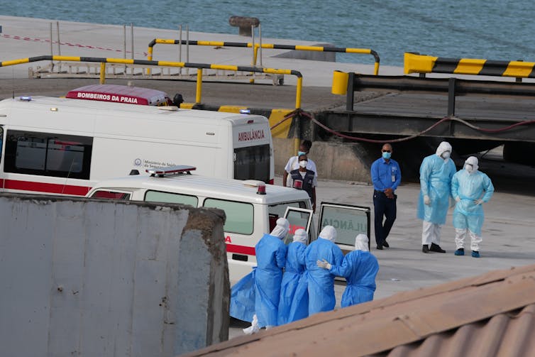 Medical staff in blue gowns on the quay of a seaport, with emergency vehicles around them