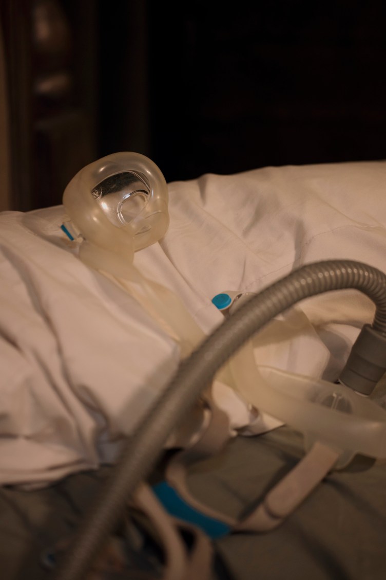A medical CPAP machine mask and tubing rest on top of white bed linens.