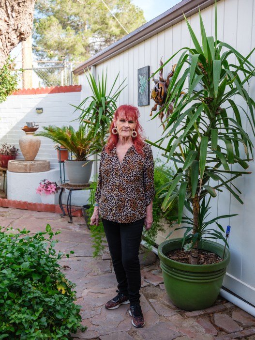 An older woman with red hair and large earrings, wearing a leopard print button-up shirt, black pants and tennis shoes, stands next to tall desert plants in the yard of a house.