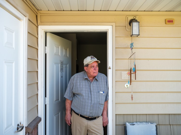 An older man wearing a white baseball hat, blue button-down shirt and khaki pants stands in the doorway to a house with a white door and yellow siding.