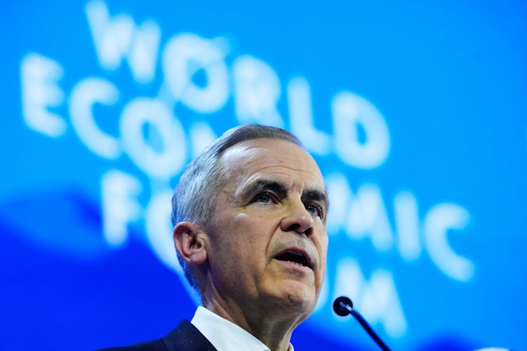 a man with short grey hair speaks in front of a large blue illuminated banner that reads world economic forum