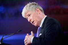 A man with white hair speaks from behind a microphone.