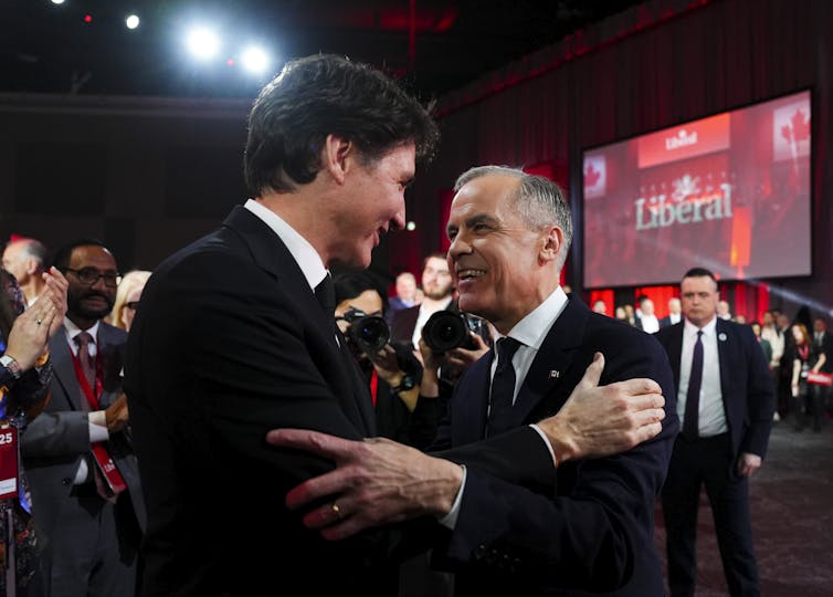 Two men — one younger and taller with dark hair, the other with short grey hair — embrace.