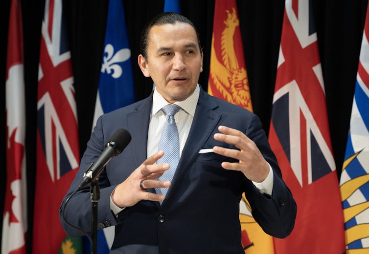 An Ojibwe man with dark hair pulled back into a short pony tail, wearing a suit, speaks from behind a podium