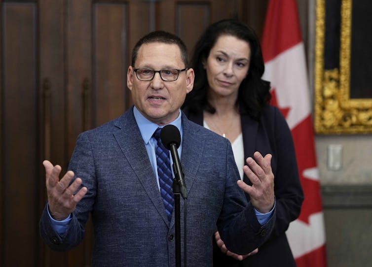 A middle-aged white man in a suit and glasses speaks from behind a microphone
