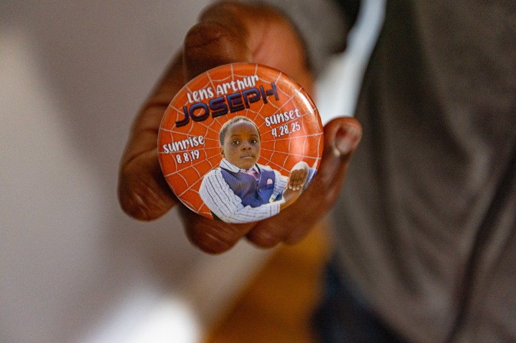 A man holds a button that has a photograph of a young child on it and the words, “Lens Arthur Joseph. Sunrise 8.8.19. Sunset 4.28.25.”