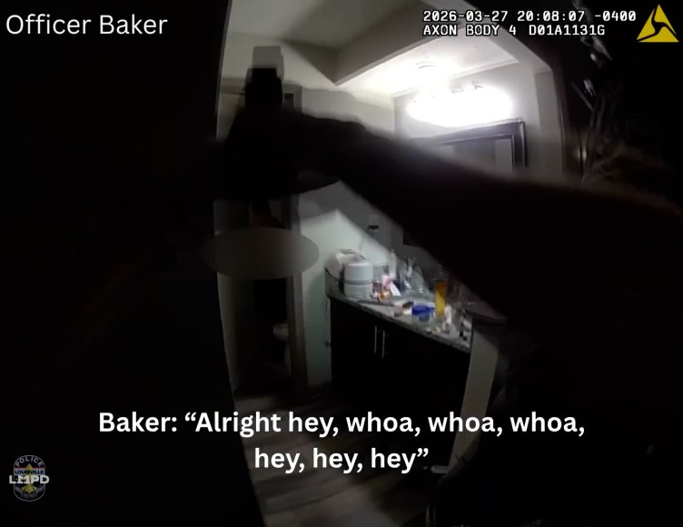 A body camera shows a police officer aiming a gun and flashlight into a bathroom as he looks at the doorway. The screen includes subtitles at the bottom reading, "Baker: 'Alright hey, whoa, whoa, whoa, hey, hey, hey.'"