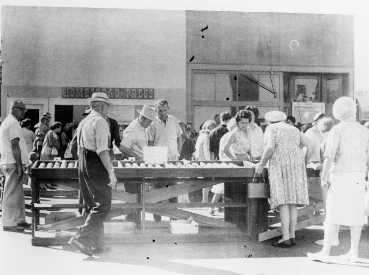 An old black-and-white photograph of a crowd of women in dresses and men in shirtsleeves looking through potatoes outside on a city street.