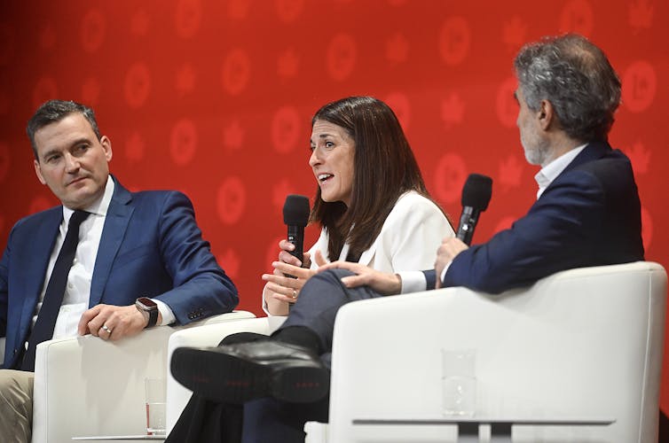 Two men and a women sit in white armchairs while speaking on a stage