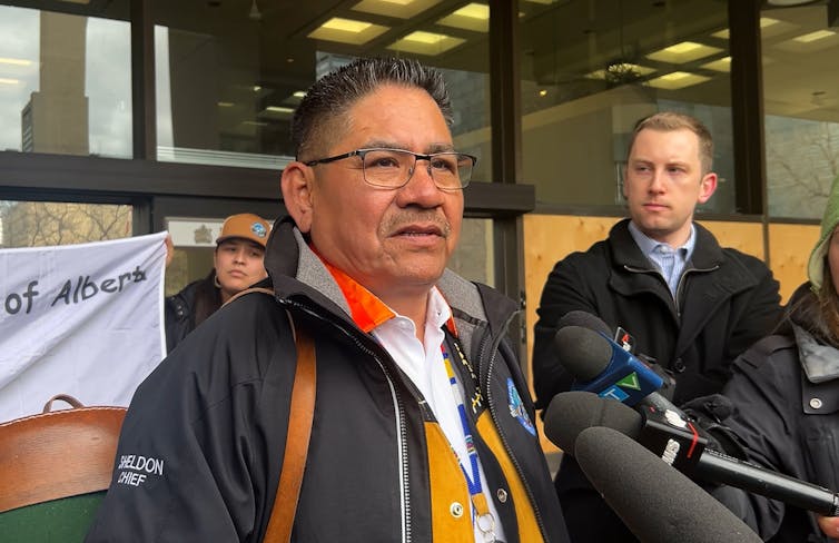 An Indigenous man wearing glasses speaks into microphones