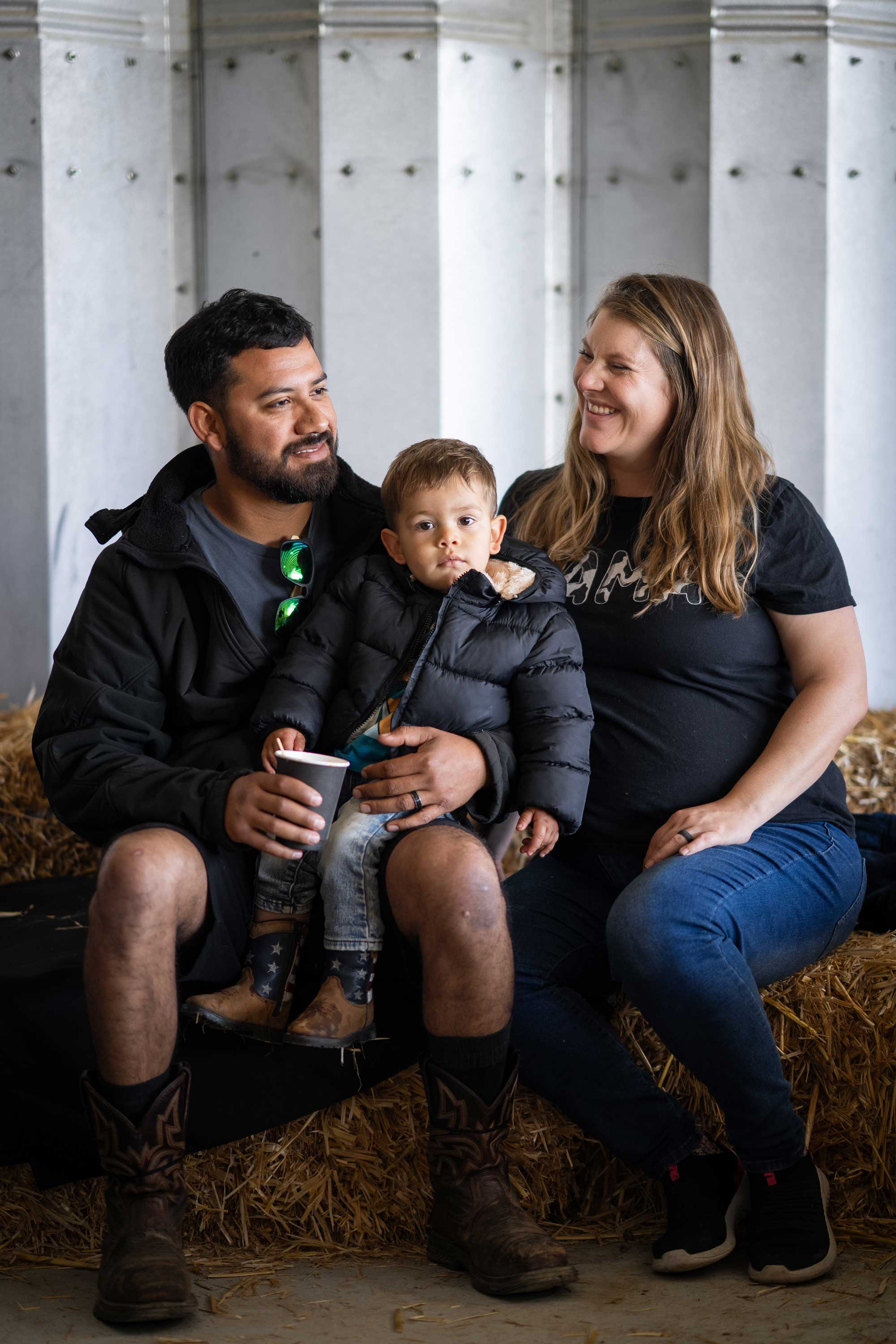 A man, a young boy sitting on his lap and a smiling woman sit together on hay bales in front of a corrugated metal wall.
