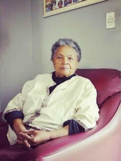 An elder African woman sits in a maroon chair and looks proudly at the camera.