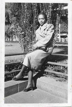 A black and white photo of a young African woman sitting cross-legged in a park in an elegant overcoat.