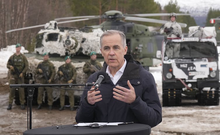 A slender man with short grey hair in front of soldiers.