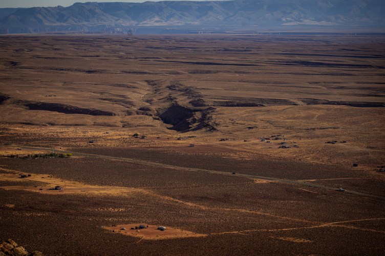 A wide, high-angle view of a vast, arid desert landscape under a hazy sky, with scattered small structures and dirt roads stretching toward a distant mountain range.