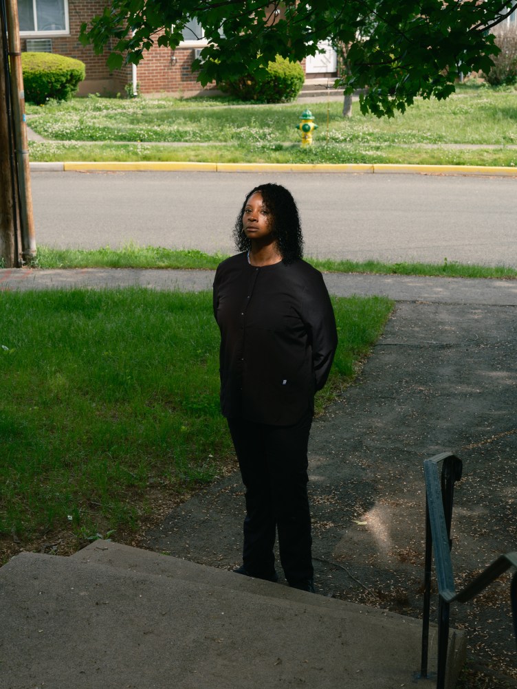 A woman stands in the walkway to a house, looking directly at the camera. She is wearing all black and her hands are tucked behind her back. A ray of light shines on her face.