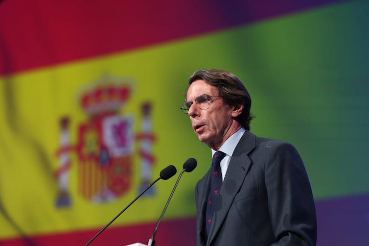 José María Aznar speaks into a microphone with the Spanish flag in the background.