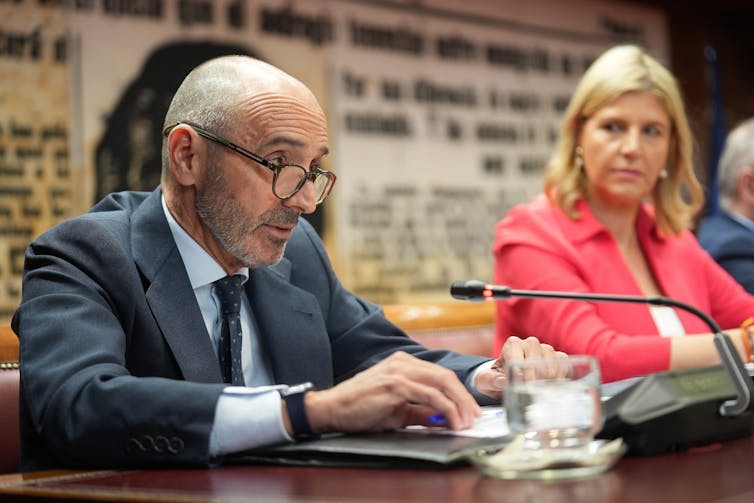 Spanish businessman Julio Martínez speaks into a microphone at a table while a woman sitting nearby watches him