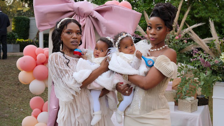 Two smartly dressed African women at a garden party with balloons, each holding a baby.