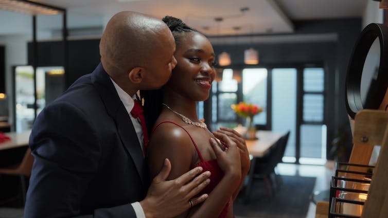 An African man in a suit kisses a young African woman from behind as she smiles into a mirror.