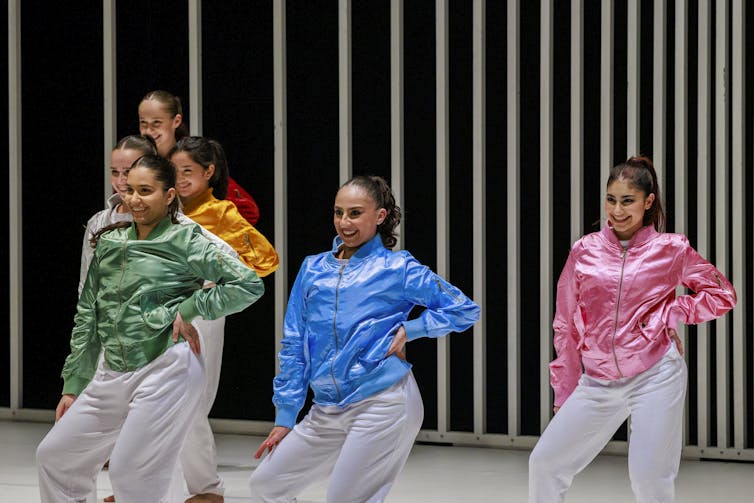 Women dancers in brighly coloured jackets pose with their hands on their hips mid-dance.