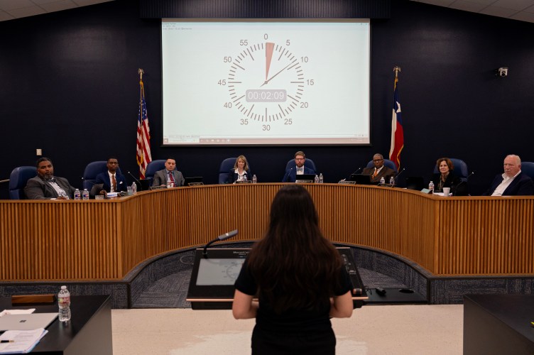 A speaker with long dark hair stands at a lectern is shown from behind, addressing a school board seated along a curved wooden dais. On the projection screen behind the board, a large digital countdown timer tracks public comment time.