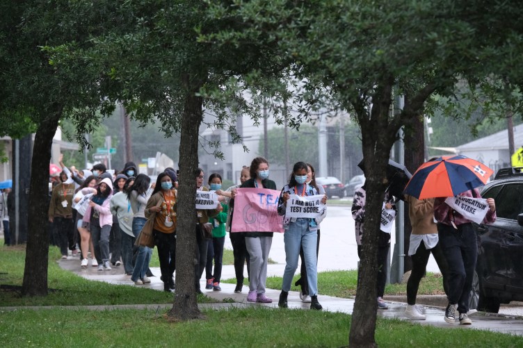 A group of students march along a rainy, tree-lined sidewalk during a protest, carrying umbrellas and signs.