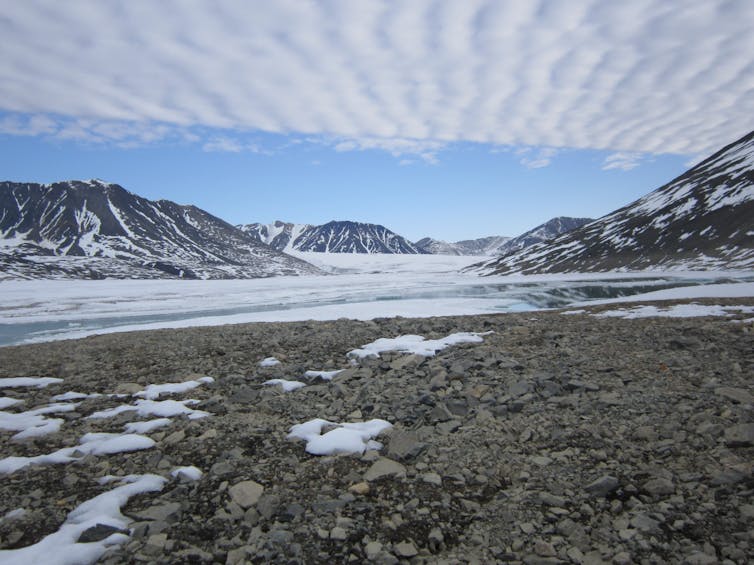 A rocky shore borders a body of water surrounded by snow-capped mountains