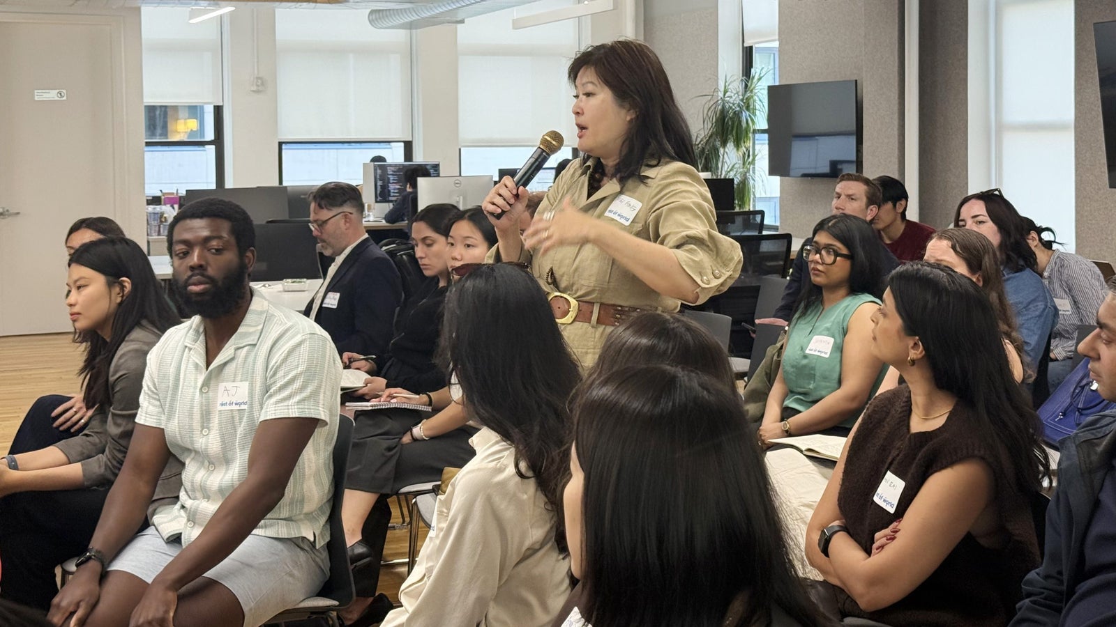 A woman in a beige outfit speaks into a microphone while seated audience members listen attentively in a modern office space.