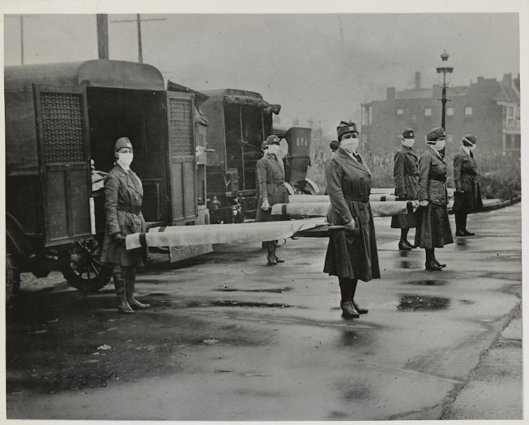 Mark-wearing nurses holding stretchers in front of military ambulances in a black and white photo from the 1918 flu pndemic
