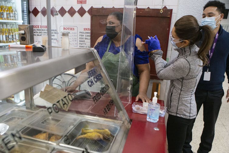 A woman in a face mask administers a shot to a woman standing near a fast-food counter