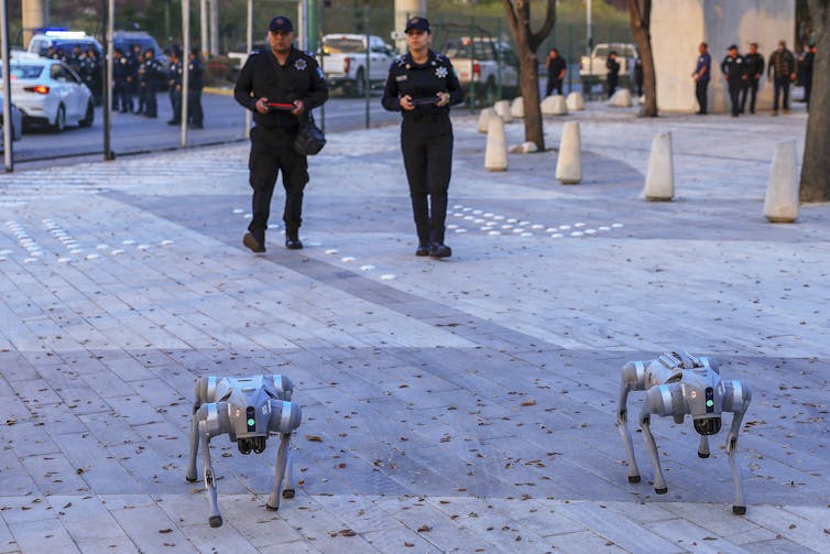 two people wearing uniforms walk behind two robotic dogs